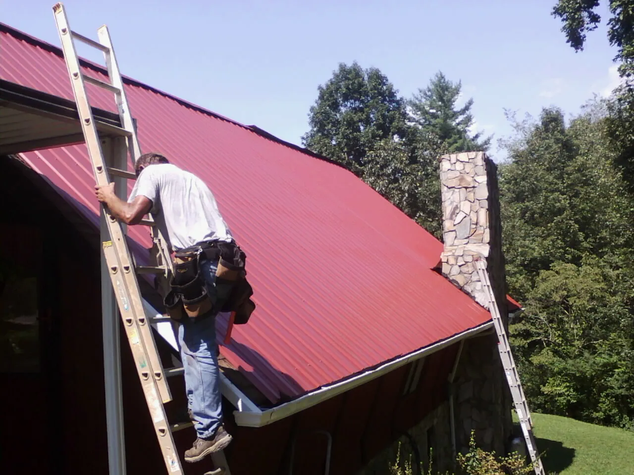 Roofer on a ladder beside a red metal roof and stone chimney — Soffit & Fascia Repair by Roofing Brothers in Carmichael